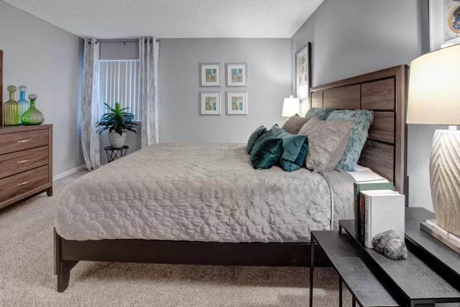 Modern bedroom with a queen bed, wooden headboard, light grey comforter, teal pillows, dresser, and framed art.