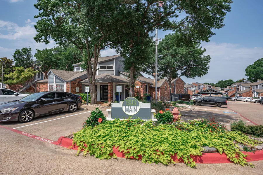 Entrance to an apartment complex featuring a sign with "MAIN," green landscaping, parked cars, and multi-story buildings.