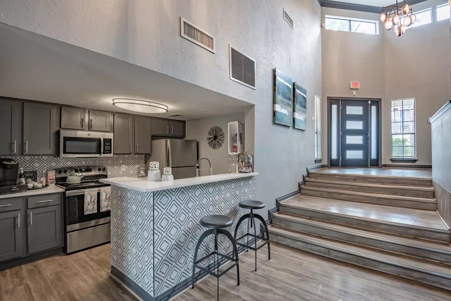 Modern common area with a gray kitchen, breakfast bar with stools, and a grand entryway with a staircase leading to double doors.