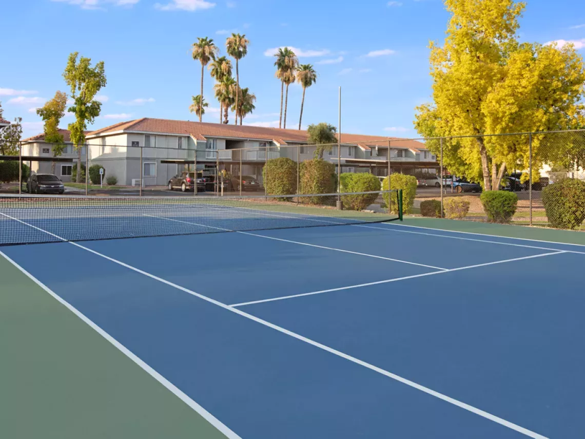 Blue and green tennis court with net, apartment buildings and palm trees in the background under a clear sky.
