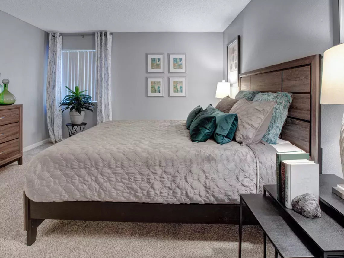 Modern bedroom with a queen bed, wooden headboard, light grey comforter, teal pillows, dresser, and framed art.