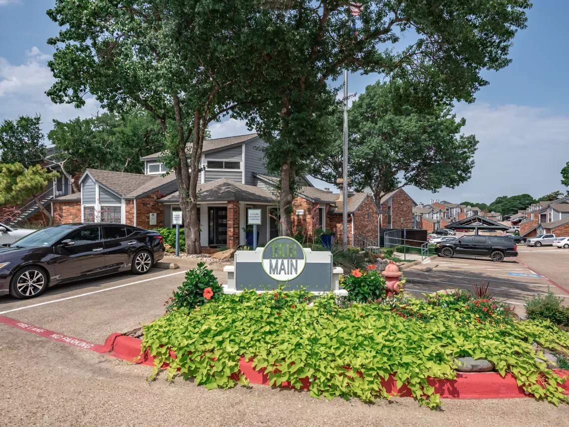 Entrance to an apartment complex featuring a sign with "MAIN," green landscaping, parked cars, and multi-story buildings.