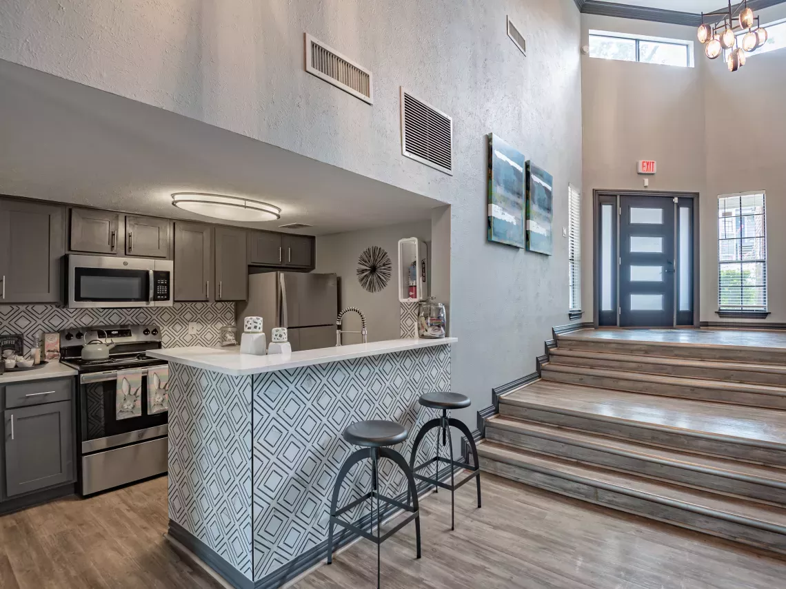 Modern common area with a gray kitchen, breakfast bar with stools, and a grand entryway with a staircase leading to double doors.