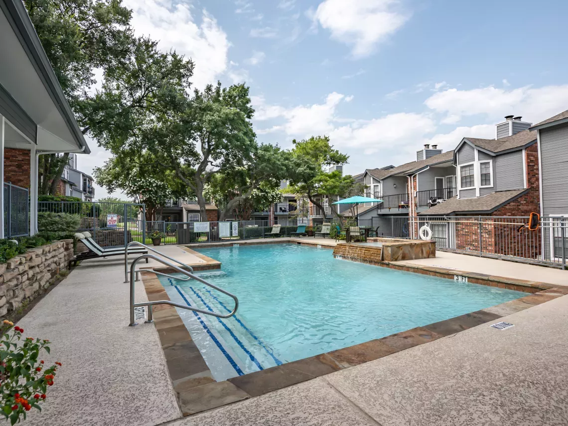 Outdoor swimming pool area with lounge chairs, a hot tub, and modern apartment buildings under a clear sky.