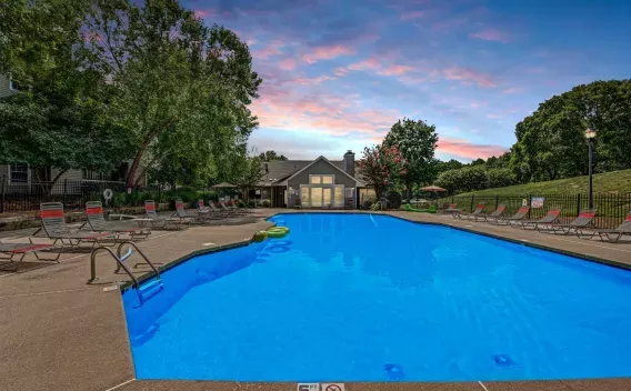 Resort-style swimming pool at 200 Braehill Apartment Community, featuring lounge chairs along the poolside, surrounded by lush greenery and trees, with a clubhouse in the background under a vibrant sunset sky."