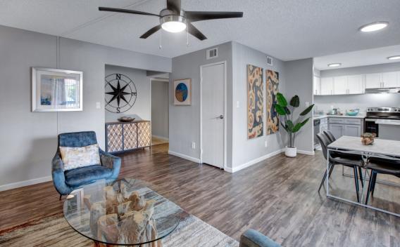 Modern open-concept living room with blue armchair, glass coffee table, dining area, and white kitchen.