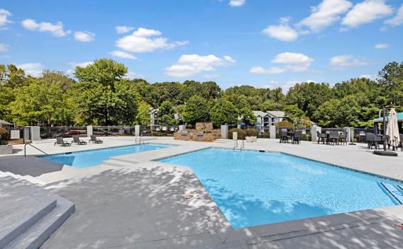 Outdoor swimming pool area with two pools, lounge chairs, and umbrellas under a bright blue sky, surrounded by green trees.