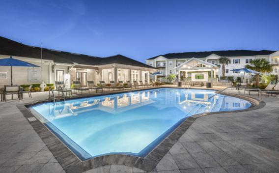Twilight view of a large, lit swimming pool surrounded by a paved patio with lounge chairs and a modern building.