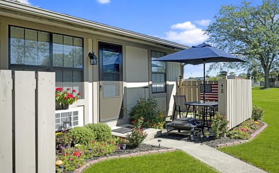 Exterior of tan two-story apartment building with two ground-level patios, lush green lawn, flower beds, and blue umbrella.