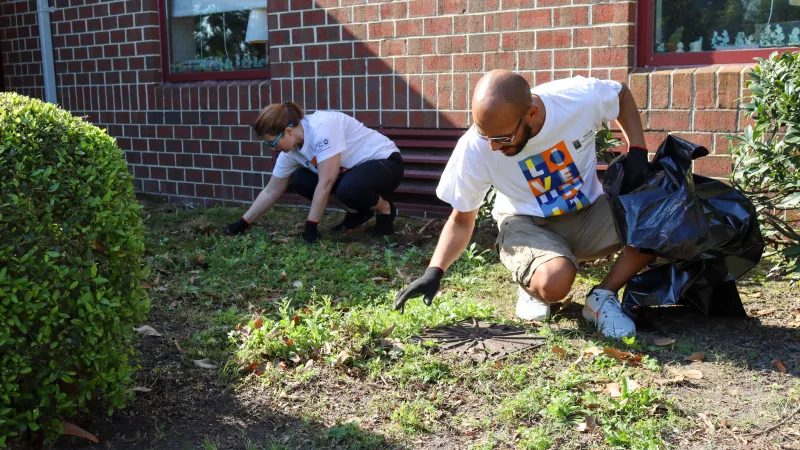 Two people in gloves pick up leaves and debris from a grassy area beside a brick building, one holding a black trash bag.