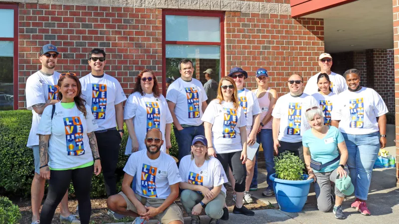 Group photo of smiling volunteers wearing white shirts with colorful logos, standing outdoors in front of a brick building.