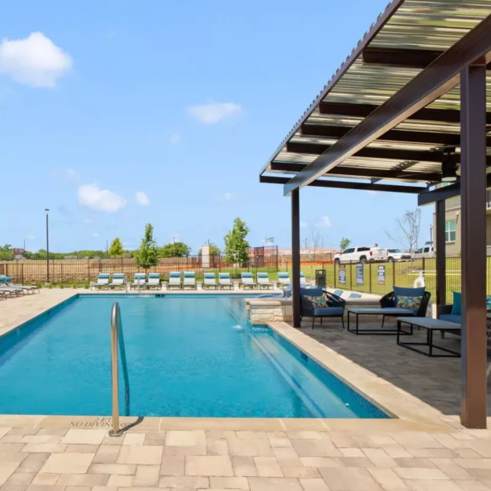 A large outdoor pool area with lounge chairs, a shaded seating area, and a modern apartment building in the background.