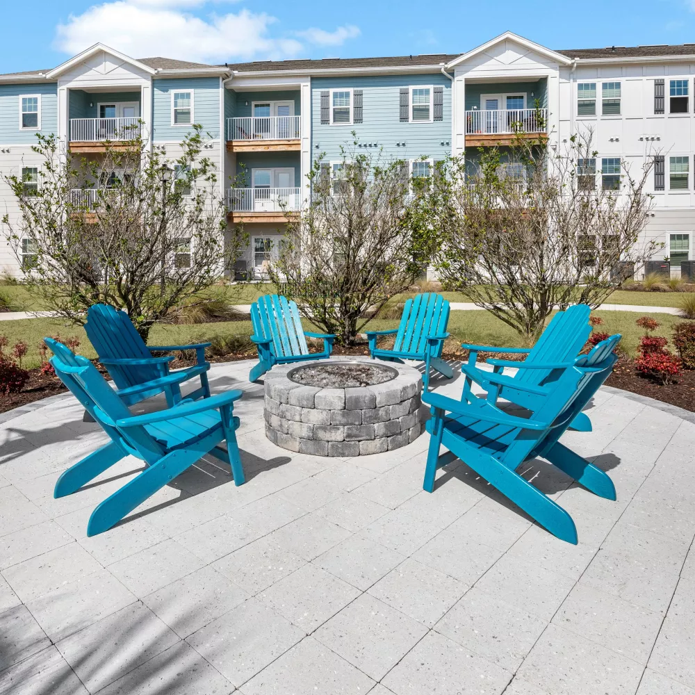 Outdoor common area with a stone fire pit, five bright blue Adirondack chairs on a patio, and a modern apartment building.