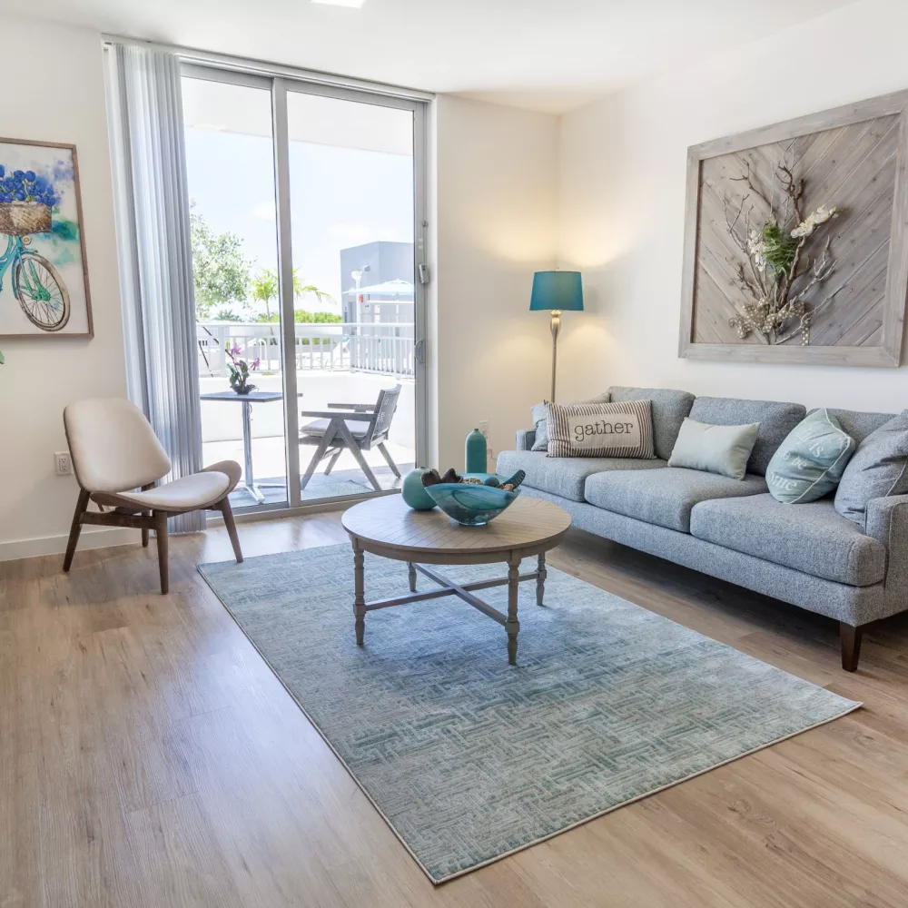 Modern living room with a light blue rug, gray sofa, wood coffee table, TV, and glass door to a balcony.