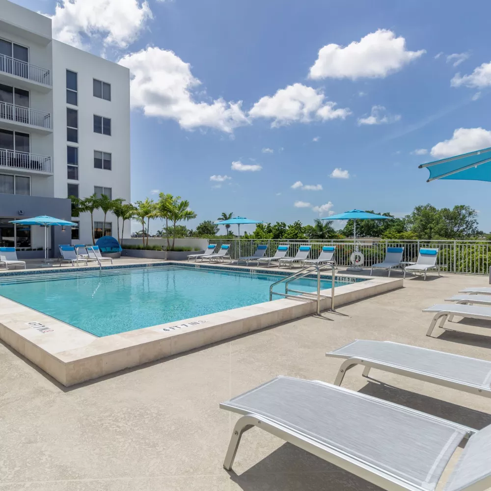 Outdoor swimming pool area with blue lounge chairs, umbrellas, and a modern white apartment building under a sunny sky.