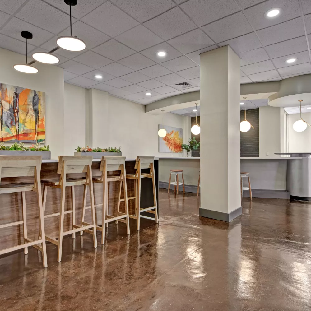 Modern resident lounge area featuring high-top seating with wooden barstools, pendant lighting, vibrant abstract artwork, and polished concrete floors.