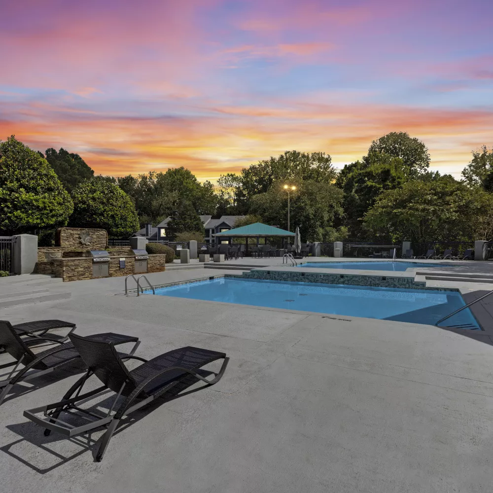 Vibrant sunset over an outdoor swimming pool area with lounge chairs on a concrete deck and lush trees.