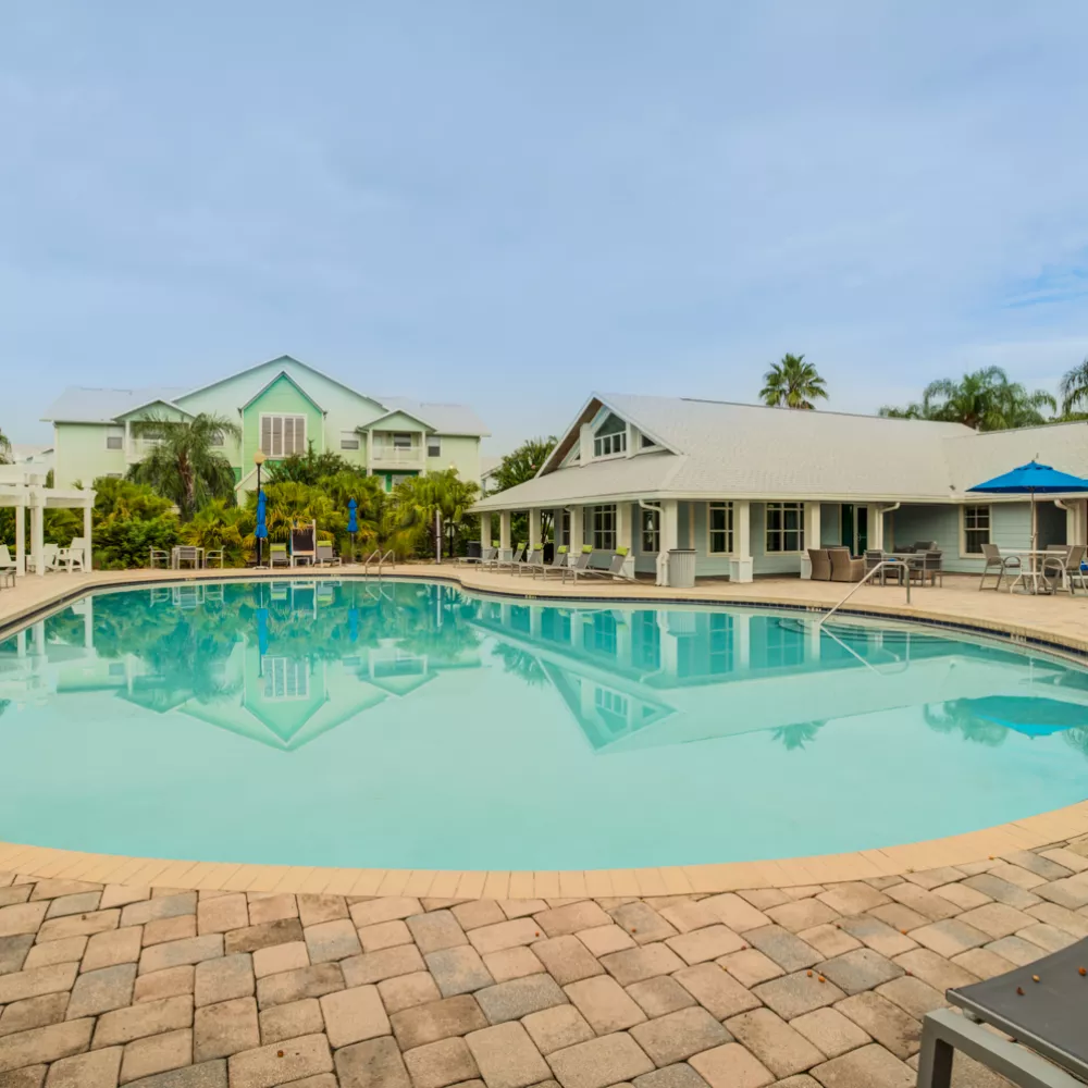 Large outdoor swimming pool with paved deck, resort buildings, palm trees, and lounge chairs under a partly cloudy sky.