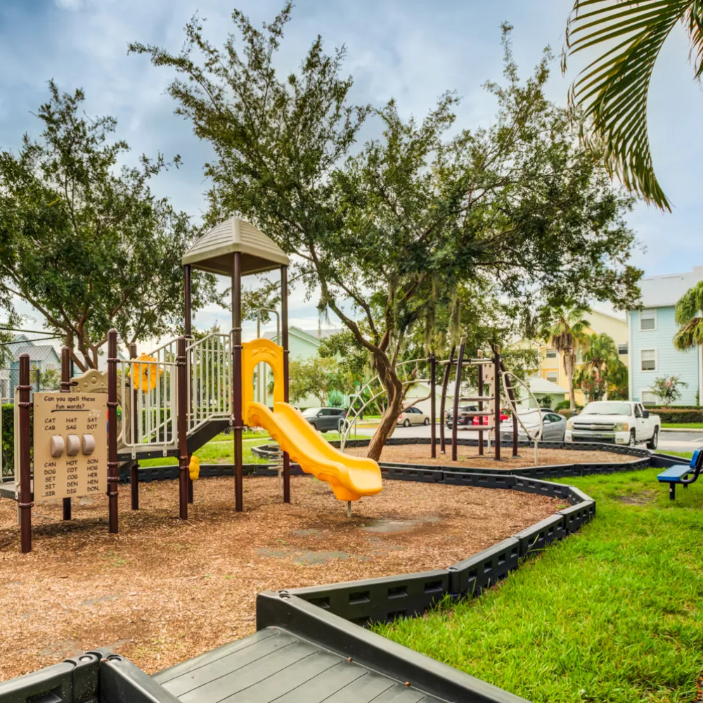 Photograph of an outdoor playground with a yellow slide, climbing structures, wood chips, trees, and residential buildings.