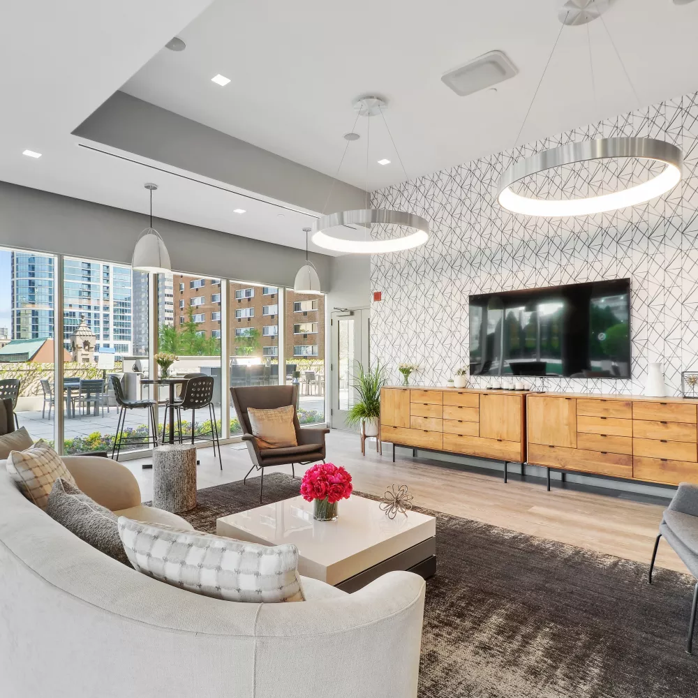 Spacious clubroom featuring a media wall with a flat-screen TV and wooden cabinetry. Large windows fill the room with natural light, highlighting the geometric accent wall and modern furniture.