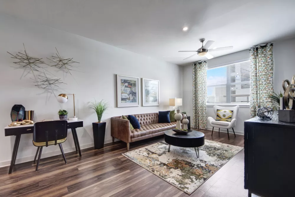 Modern living room with a brown leather sofa, round coffee table, desk, and a large window with patterned curtains.