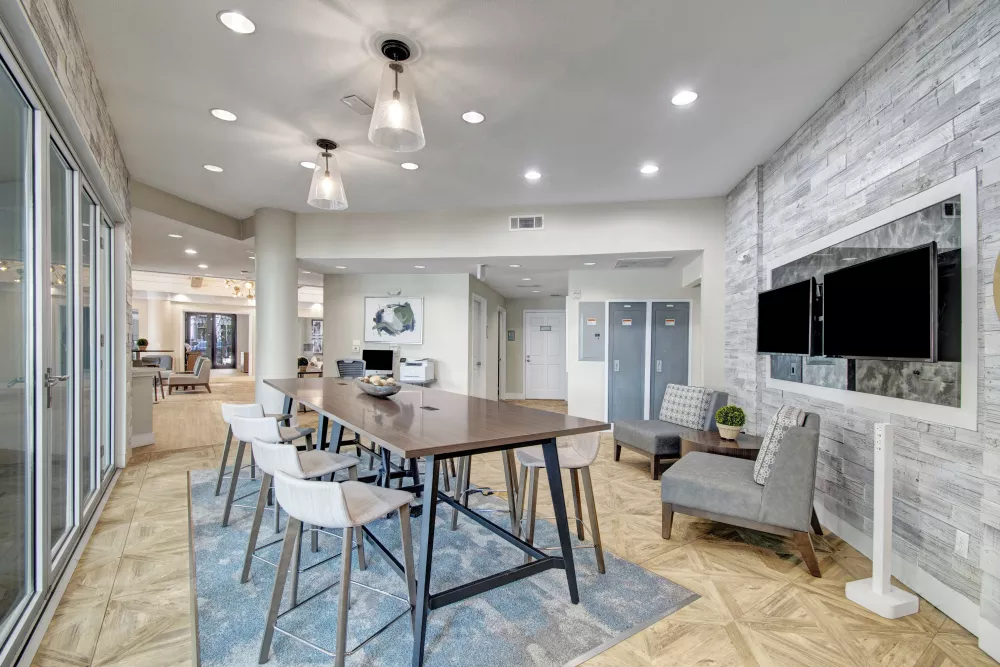 Bright, modern lounge with a long wooden table, white stools, blue rug, armchairs, and wall-mounted TVs.
