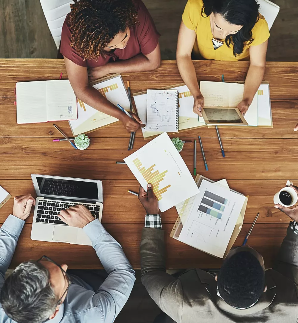 Top-down view of a diverse group of professionals collaborating around a wooden table in a modern office setting. They are analyzing data, discussing reports with charts and graphs, using a laptop and a tablet, and taking notes. The workspace is filled with pens, notebooks, and small potted plants, reflecting a dynamic and productive teamwork environment.