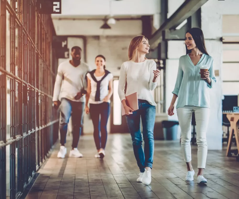 **Alt text:** "Group of four young professionals walking and talking in a modern, open-concept office space with industrial-style design. Two women in the foreground are engaged in conversation, one holding a laptop and the other carrying a coffee cup. Two colleagues, a man and a woman, walk behind them, all dressed casually. The office features exposed metal railings, large windows, and wooden floors, creating a collaborative and dynamic work environment."