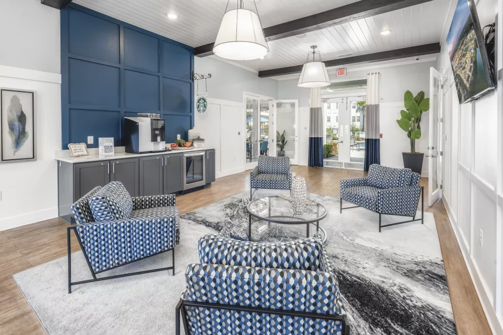 Stylish community lounge featuring a blue paneled wall, coffee station, patterned chairs, and a large rug.