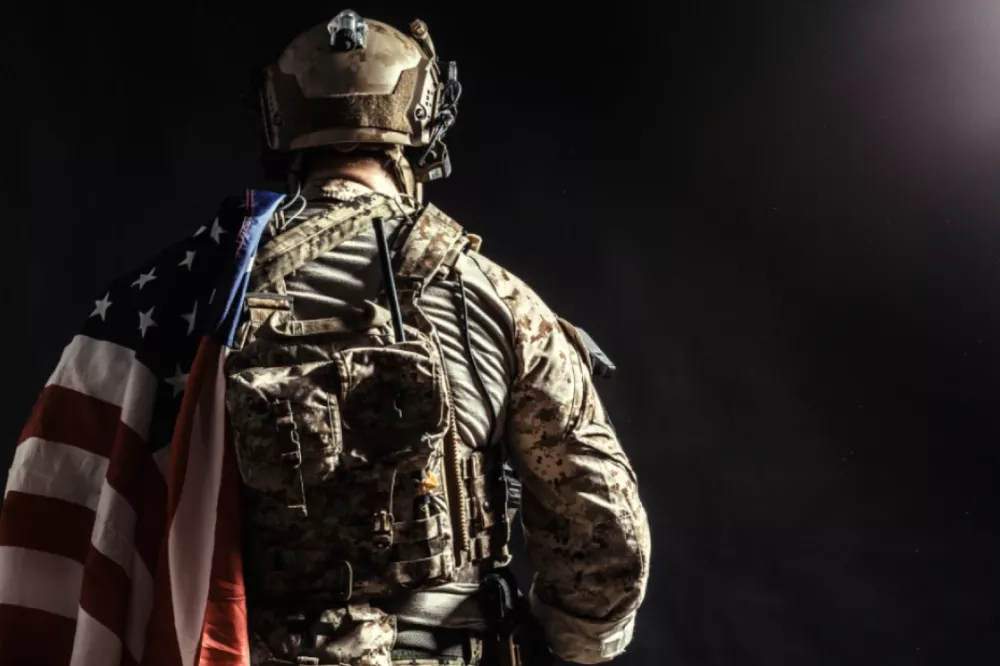 A soldier wearing tactical gear with an American flag draped over their shoulder, standing in a dimly lit setting.