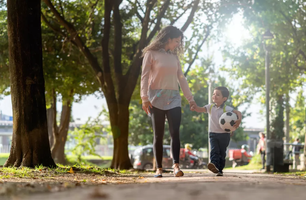 A person and a child playing with a football in an outdoor park setting, surrounded by grass and trees. They are wearing casual clothing and footwear, enjoying the sunny day together.