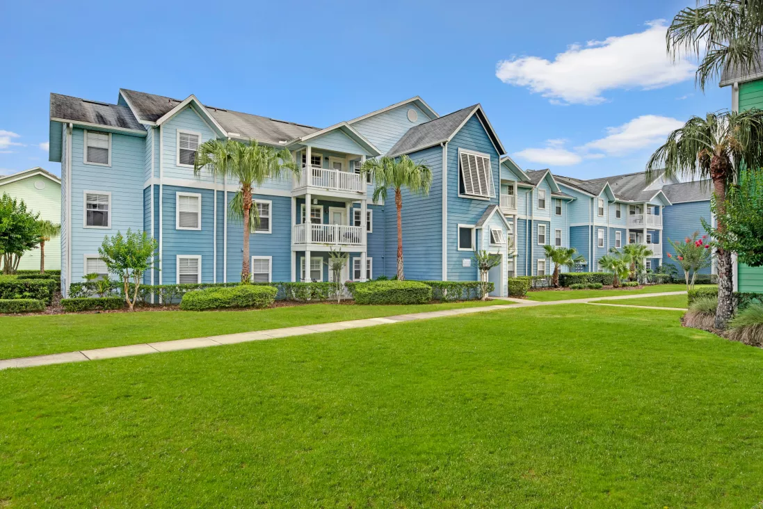 Blue multi-story apartment building with white balconies, surrounded by green lawns and palm trees.