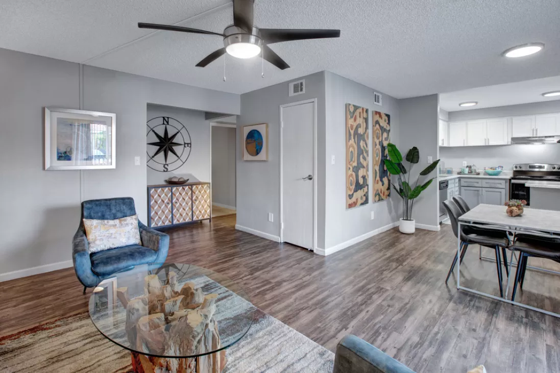 Modern open-concept living room with blue armchair, glass coffee table, dining area, and white kitchen.