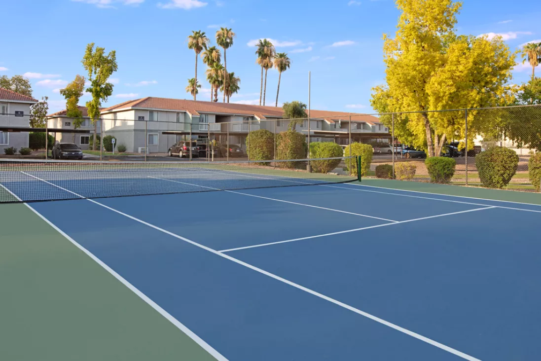 Empty blue and green tennis court with apartment buildings and palm trees under a clear sky.