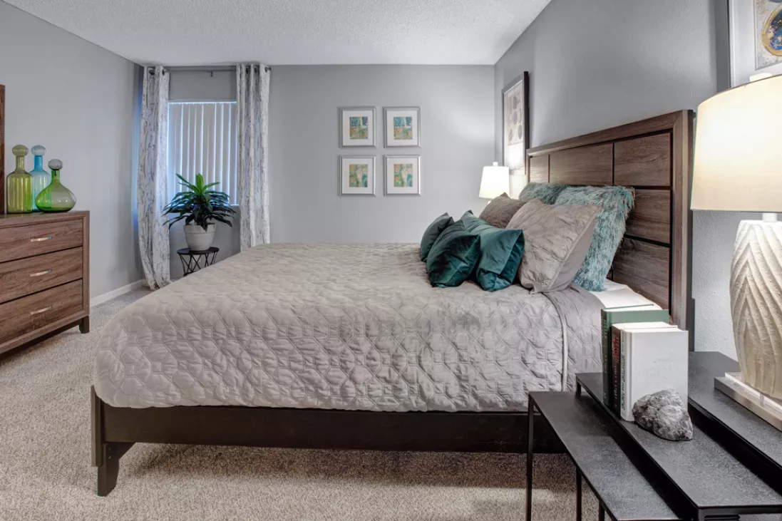 Modern bedroom with a queen bed, wooden headboard, light grey comforter, teal pillows, dresser, and framed art.