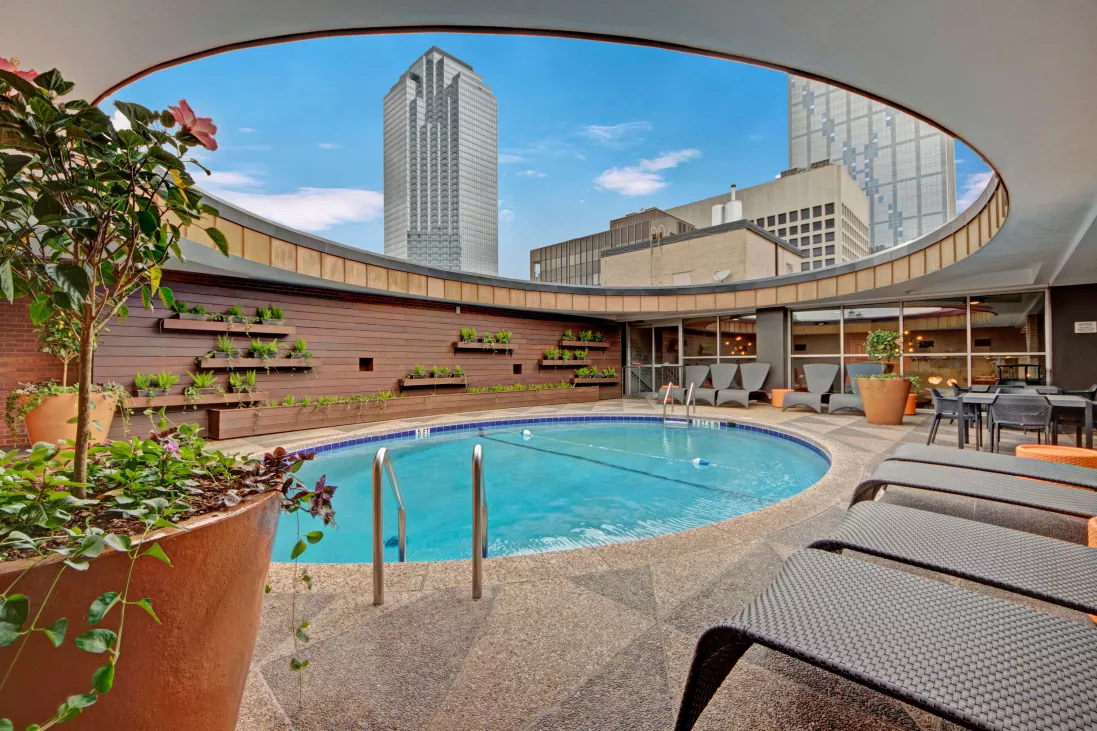 Unique circular rooftop pool surrounded by modern lounge seating and greenery, with views of downtown skyscrapers and urban skyline.