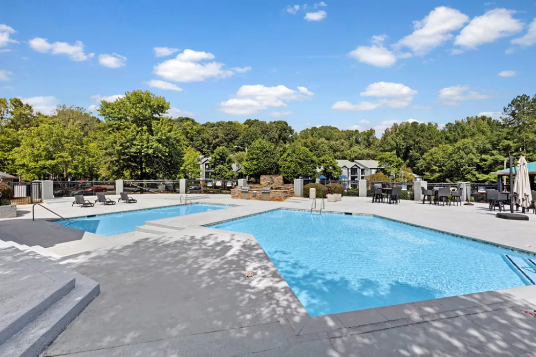 Outdoor swimming pool area with two pools, lounge chairs, and umbrellas under a bright blue sky, surrounded by green trees.