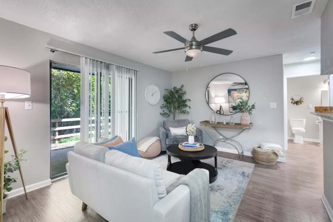 Stylish apartment living room featuring a white sofa, a gray accent chair, a round mirror, and a balcony overlooking trees.