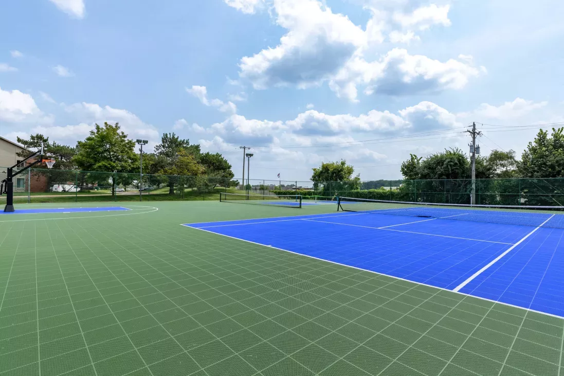 Outdoor sports court with green and blue sections under a blue sky with clouds. A net is visible.