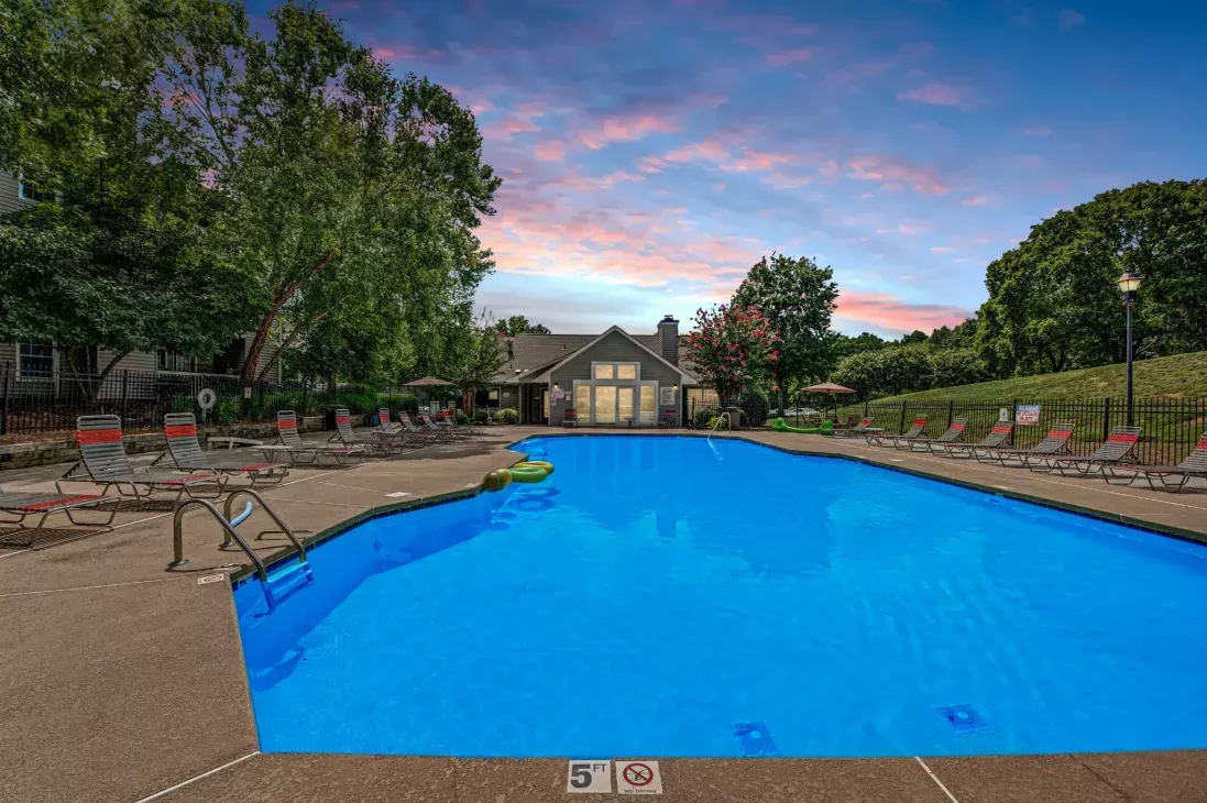 Resort-style swimming pool at 200 Braehill Apartment Community, featuring lounge chairs along the poolside, surrounded by lush greenery and trees, with a clubhouse in the background under a vibrant sunset sky."