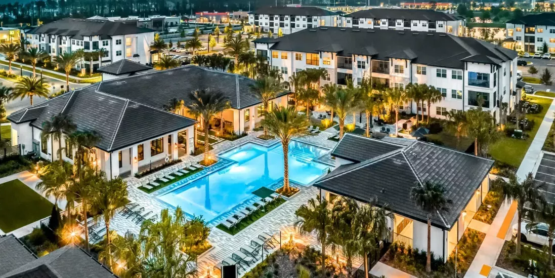 An aerial night view of a vibrant residential community with a stunning illuminated pool, palm trees, and surrounding apartment buildings.