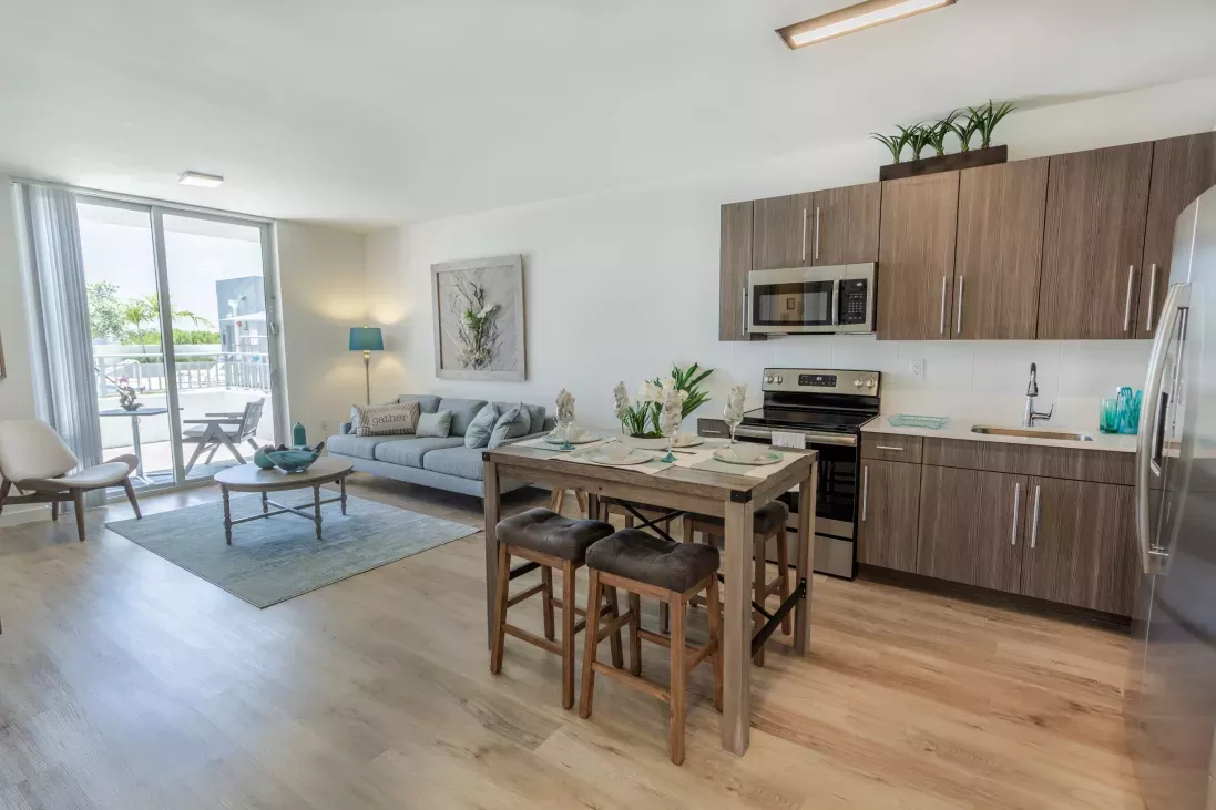Bright, open-plan living room with gray sofa, blue rug, and a modern kitchen with wood cabinets and island.
