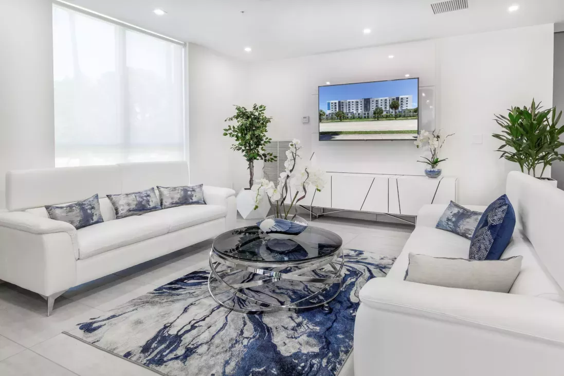 Bright, modern waiting room with two white sofas, a round coffee table, a blue abstract rug, and a TV displaying a building.