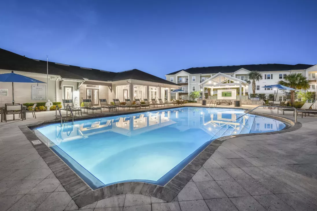 Twilight view of a large, lit swimming pool surrounded by a paved patio with lounge chairs and a modern building.
