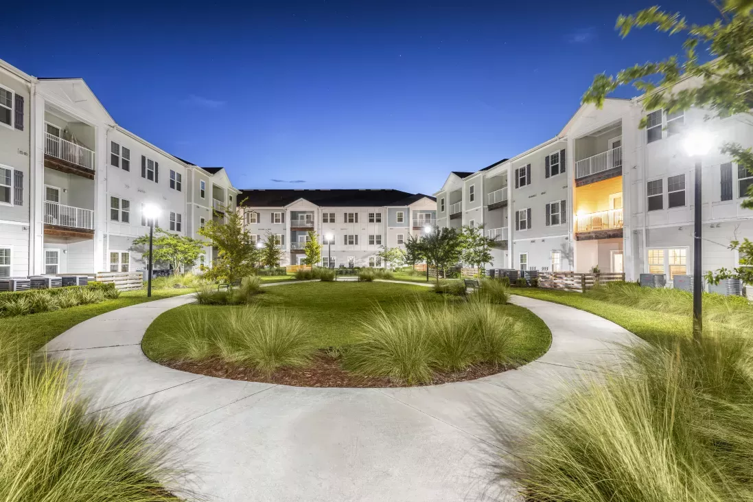 Illuminated courtyard of a modern apartment complex at night, featuring white buildings, curved paths, and lush greenery.