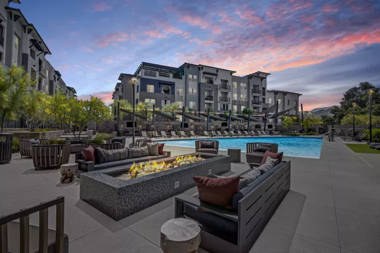Modern apartment complex exterior at dusk with a lit fire pit, patio seating, and a swimming pool.